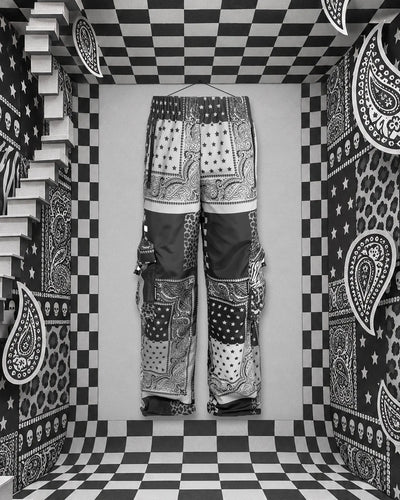 Το παντελόνι cargo Romanticbandana, patchwork από μαύρες, λευκές και γκρι μπαντάνες, κρεμασμένο με ένα νήμα από ένα καρφί στον πίσω τοίχο ενός χάρτινου δωματίου σε λευκό και μαύρο καρό. Αριστερά μια λευκή σκάλα, στους τοίχους paisley, αστέρια, ζεβρέ, κηλίδες και μικρά κρανία.