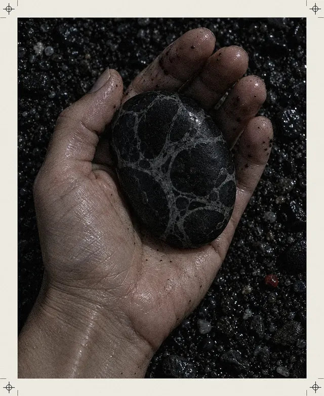 Una mano bagnata tiene nel palmo un ciottolo nero con venature chiare come la stampa Strand, sulla ghiaia scura, e accanto c'è un sassolino rosso.